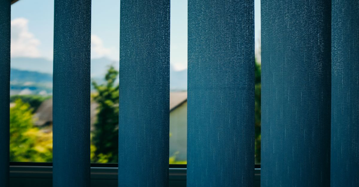 Through blue blinds view on green trees growing in yard of home located in suburb