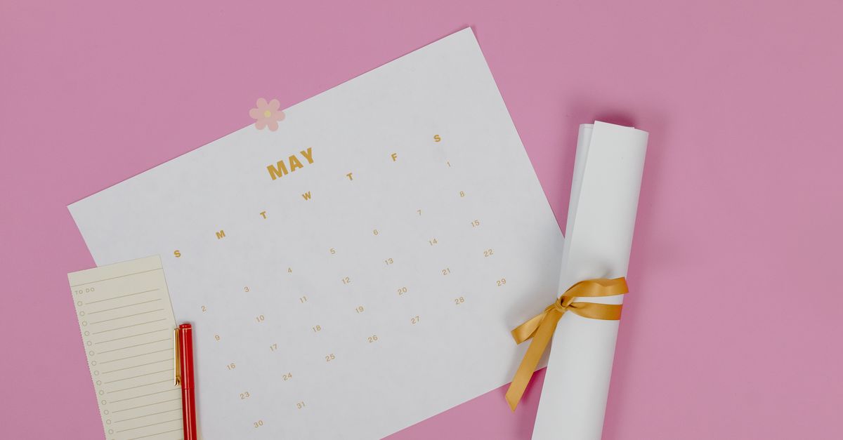 Flat lay of a May calendar with a pen and paper roll on pink background.